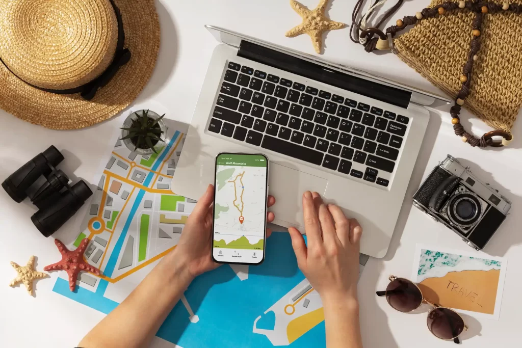 Startups TravelTech. Mesa con ordenador portátil, móvil en el que se ve un mapa y alrededor podemos ver accesorios de vacaciones, un mapa físico, sombrero de playa, gafas de sol, cámara de fotos y prismáticos